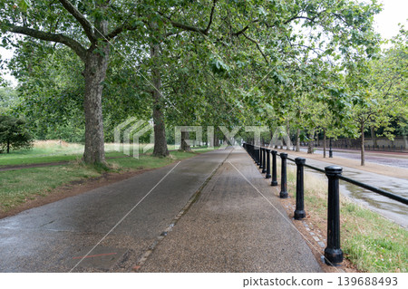 Long pedestrian walkway stretches through a tree lined urban park with wet pavement, cloudy weather, and peaceful outdoor scenery. Long pedestrian walkway stretches through a tree lined urban park with wet pavement, cloudy weather, and peaceful outdoor scenery. 139688493