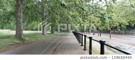 Long pedestrian walkway stretches through a tree lined urban park with wet pavement, cloudy weather, and peaceful outdoor scenery. 139688494