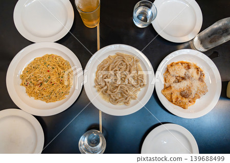Italian pasta dishes with creamy squid noodles and ravioli are arranged across a dark restaurant table with white plates and drinks. Italian pasta dishes with creamy squid noodles and ravioli are arranged across a dark restaurant table with white plates and drinks. 139688499