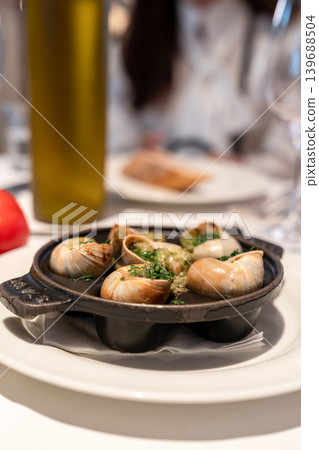 Cooked escargot in shells is topped with garlic herb butter on a black pan. White plates and drinks appear blurred in the restaurant background. Cooked escargot in shells is topped with garlic herb butter on a black pan. White plates and drinks appear blurred in the restaurant background. 139688504