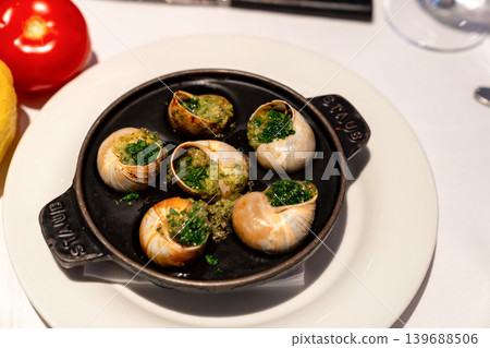 Cooked escargot in shells is topped with garlic herb butter on a black pan. White plates and drinks appear blurred in the restaurant background. 139688506