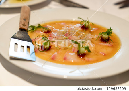 Thinly sliced fish served with citrus dressing, garnished with microgreens and decorative sauce dots. Plate and fork visible on white table. Thinly sliced fish served with citrus dressing, garnished with microgreens and decorative sauce dots. Plate and fork visible on white table. 139688509