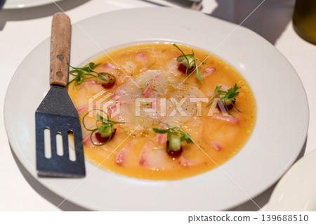 Thinly sliced fish served with citrus dressing, garnished with microgreens and decorative sauce dots. Plate and fork visible on white table. Thinly sliced fish served with citrus dressing, garnished with microgreens and decorative sauce dots. Plate and fork visible on white table. 139688510