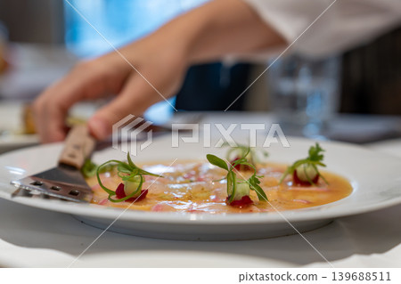 Thinly sliced fish served with citrus dressing, garnished with microgreens and decorative sauce dots. Plate and fork visible on white table. Thinly sliced fish served with citrus dressing, garnished with microgreens and decorative sauce dots. Plate and fork visible on white table. 139688511