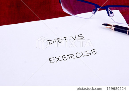 A white sheet of paper displays the handwritten words DIET VS EXERCISE. A fountain pen and blue reading glasses rest on the paper, set against a red background A white sheet of paper displays the handwritten words DIET VS EXERCISE. A fountain pen and blue reading glasses rest on the paper, set against a red background 139689224