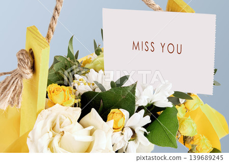 Close up of white and yellow roses with daisies in a rustic yellow wooden box. A white card with Miss You in brown text rests above the flowers, conveying longing Close up of white and yellow roses with daisies in a rustic yellow wooden box. A white card with Miss You in brown text rests above the flowers, conveying longing 139689245