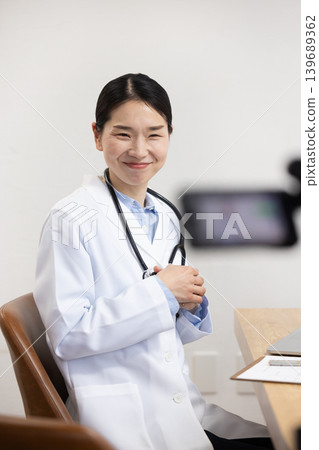 A Japanese female doctor in a white coat speaks with hand gestures in front of a video camera. A Japanese female doctor in a white coat speaks with hand gestures in front of a video camera. 139689362