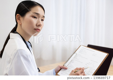 A Japanese female doctor in a white coat speaks while holding a medical chart. A Japanese female doctor in a white coat speaks while holding a medical chart. 139689717