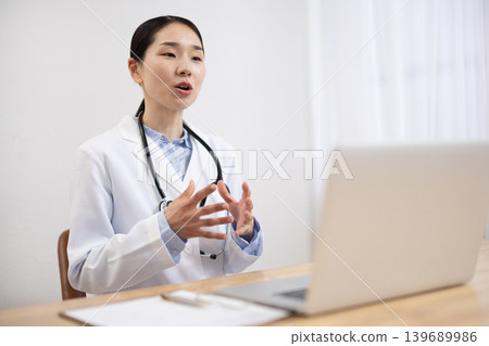 Japanese female doctor in a white coat making an online call on her computer Japanese female doctor in a white coat making an online call on her computer 139689986