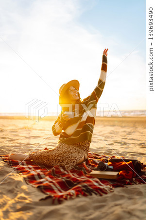 Happy woman in a stylish sweater sits at picnic on the beach drinks a hot drink from a thermos Happy woman in a stylish sweater sits at picnic on the beach drinks a hot drink from a thermos 139693490