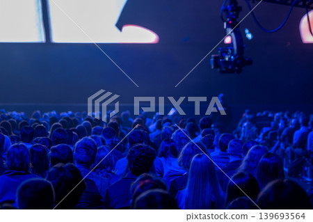 Audience watching conference presentation in dark auditorium with blue lighting and camera crane filming Audience watching conference presentation in dark auditorium with blue lighting and camera crane filming 139693564