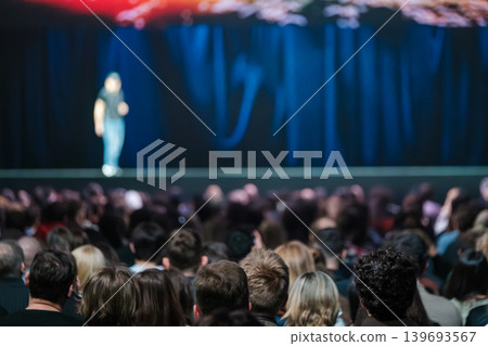 Audience watching keynote speaker on stage during conference in dark auditorium, blurred focus 139693567