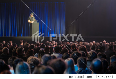 Large audience watching presenter on stage during conference presentation with blue backdrop and spotlight Large audience watching presenter on stage during conference presentation with blue backdrop and spotlight 139693571