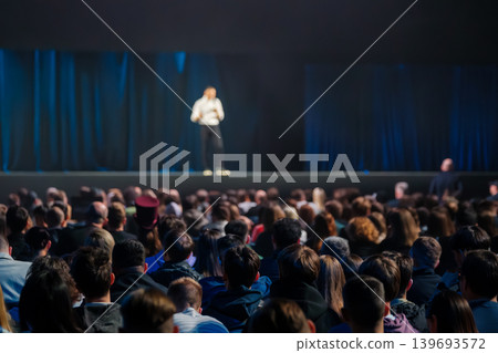 Large audience watching speaker on stage during conference presentation with dim blue curtains 139693572