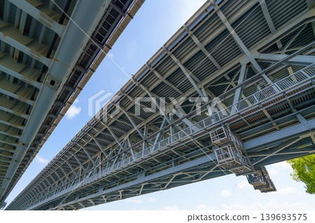 Low-angle view of the bridge's underside: complex steel truss geometric structure and blue sky engineering photograph. 139693575