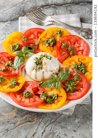 burrata cheese served with sliced red and yellow tomatoes, fresh basil leaves, and green pesto sauce on a white plate with two forks. Vertical burrata cheese served with sliced red and yellow tomatoes, fresh basil leaves, and green pesto sauce on a white plate with two forks. Vertical 139694192
