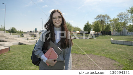A hand-held shot of a young female student in casual clothes, wearing glasses and a backpack, holding a notebook and laptop in her hands, walking through an urban park on a summer day. A hand-held shot of a young female student in casual clothes, wearing glasses and a backpack, holding a notebook and laptop in her hands, walking through an urban park on a summer day. 139694321