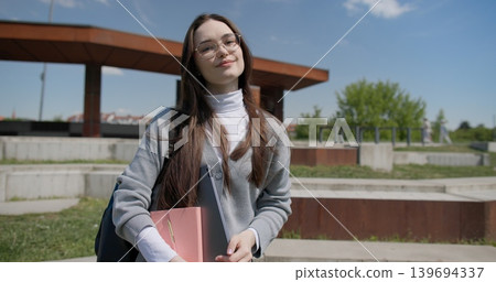 A young beautiful brunette student girl in casual clothes wearing glasses with a backpack and a laptop and a notepad in her hands stands outside against the backdrop of a city urban park A young beautiful brunette student girl in casual clothes wearing glasses with a backpack and a laptop and a notepad in her hands stands outside against the backdrop of a city urban park 139694337