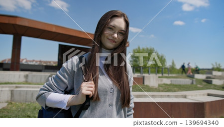A young beautiful brunette student girl in casual clothes with glasses and a backpack stands outside on a sunny summer day against the backdrop of a city urban park 139694340