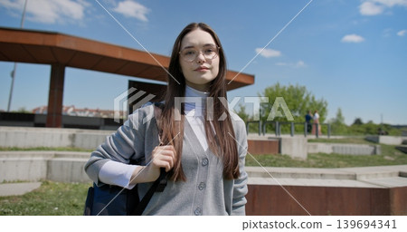 A young beautiful brunette student girl in casual clothes with glasses and a backpack stands outside on a sunny summer day against the backdrop of a city urban park 139694341