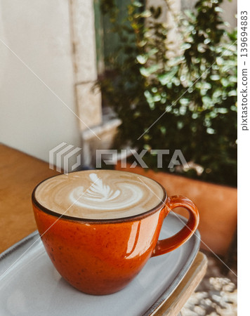 Orange cup of cappuccino with latte art on a cafe table, cozy coffee scene with soft natural light, green plants and warm atmosphere Orange cup of cappuccino with latte art on a cafe table, cozy coffee scene with soft natural light, green plants and warm atmosphere 139694883