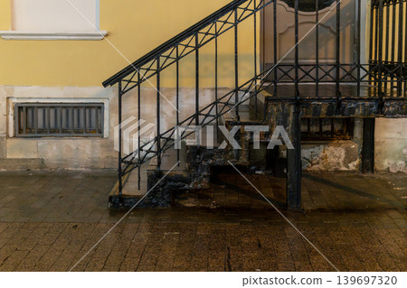 Rusted Metal Staircase With Iron Railing Against Yellow Building Facade On Wet Urban Sidewalk 139697320