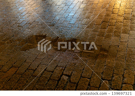 Wet Cobblestone Street With Night Puddle Reflecting City Lights And Glistening Brick Texture Wet Cobblestone Street With Night Puddle Reflecting City Lights And Glistening Brick Texture 139697321