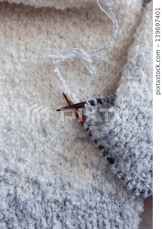White and grey boucle knitting with knitting needles. Soft white knitwear with wooden needles. Knitting pattern background. Knitting lifestyle. Cozy hobby. Warm knitwear. Craft product concept White and grey boucle knitting with knitting needles. Soft white knitwear with wooden needles. Knitting pattern background. Knitting lifestyle. Cozy hobby. Warm knitwear. Craft product concept 139697401
