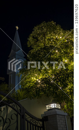 Night view of a church tower with glowing cross and clock surrounded by illuminated tropical trees and decorative lights in peaceful urban atmosphere. Night view of a church tower with glowing cross and clock surrounded by illuminated tropical trees and decorative lights in peaceful urban atmosphere. 139697620