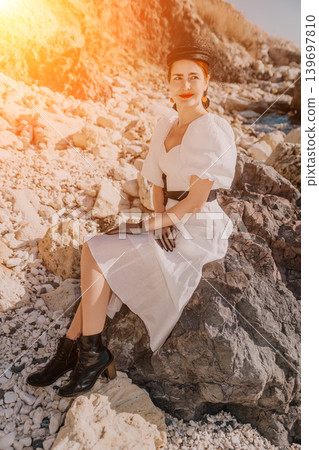 Woman white dress sunset elegant lady sitting on coastal rocks during golden hour for a summer lifestyle portrait 139697810