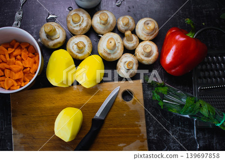 Dinner preparing process. Vegetable soup ingredients. Peeled potatoes, carrot with mushrooms and red bell pepper on the table. Raw vegetables with knife on wooden board. Healthy eating. Raw food Dinner preparing process. Vegetable soup ingredients. Peeled potatoes, carrot with mushrooms and red bell pepper on the table. Raw vegetables with knife on wooden board. Healthy eating. Raw food 139697858