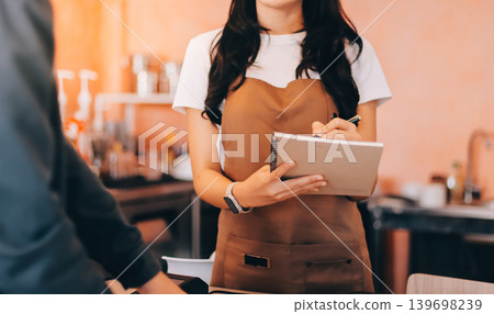 Friendly Asian female barista receives a credit card from a customer for payment. Both women are smiling in a warmly lit coffee shop with a rustic interior. Cashless payments. Small family business Friendly Asian female barista receives a credit card from a customer for payment. Both women are smiling in a warmly lit coffee shop with a rustic interior. Cashless payments. Small family business 139698239