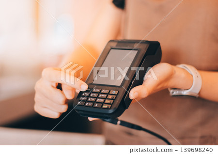 Friendly Asian female barista receives a credit card from a customer for payment. Both women are smiling in a warmly lit coffee shop with a rustic interior. Cashless payments. Small family business Friendly Asian female barista receives a credit card from a customer for payment. Both women are smiling in a warmly lit coffee shop with a rustic interior. Cashless payments. Small family business 139698240