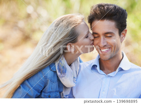 Happy couple, kiss and cheek in nature for love, bonding or support in relax for outdoor affection. Face of young woman and man smile for embrace, intimacy or romance in forest or woods together 139698895