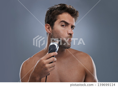 Man, shaving and machine in studio for beard grooming, skincare and portrait by grey background. Person, model and self care for hygiene, facial hair removal and electric razor for cosmetic change 139699114
