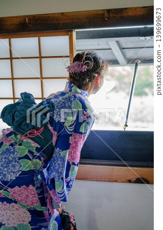 A woman in a yukata with a hydrangea pattern, seen from behind, gazing out the window of a traditional wooden building. Aichi Prefecture, Japan. A woman in a yukata with a hydrangea pattern, seen from behind, gazing out the window of a traditional wooden building. Aichi Prefecture, Japan. 139699673