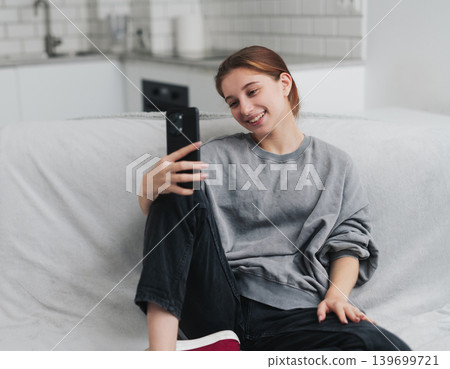 Happy, Pretty Young Girl Sitting On The Sofa At Home And Talking Via Video Call On Smartphone Happy, Pretty Young Girl Sitting On The Sofa At Home And Talking Via Video Call On Smartphone 139699721
