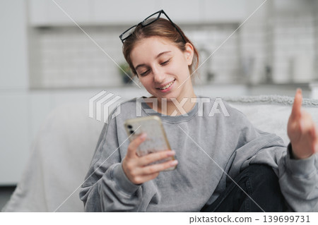 Happy Girl Sitting On The Sofa In A Light Room, Talking On A Video Call With Phone Happy Girl Sitting On The Sofa In A Light Room, Talking On A Video Call With Phone 139699731