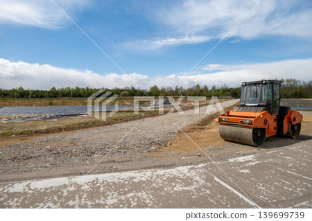 An asphalt paver works on a dirt road at a construction site under a clear sky. Nearby, there are grassy areas and materials An asphalt paver works on a dirt road at a construction site under a clear sky. Nearby, there are grassy areas and materials 139699739