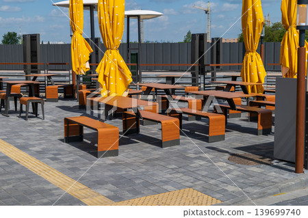 Several wooden tables and benches are set up in an outdoor area. Yellow curtains are hanging, and the sky is clear 139699740