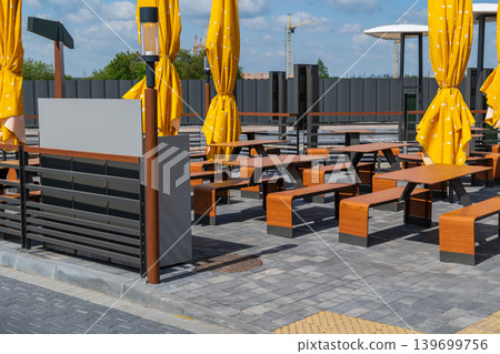 A street cafe features wooden tables and yellow umbrellas. People enjoy drinks and food in a lively atmosphere under clear blue skies 139699756