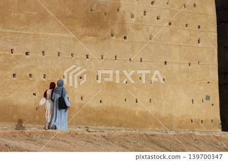 Two women walk beside a tall, textured stone wall Two women walk beside a tall, textured stone wall 139700347