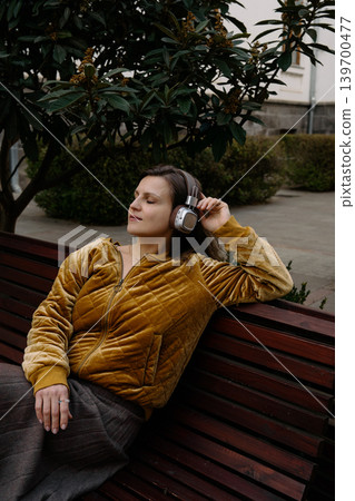 Woman sits on a bench in a park while listening to music with headphones Woman sits on a bench in a park while listening to music with headphones 139700477