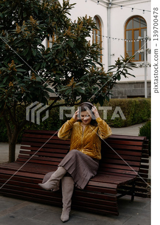 Woman sits on a bench wearing a yellow jacket and headphone in a garden setting during daytime Woman sits on a bench wearing a yellow jacket and headphone in a garden setting during daytime 139700478