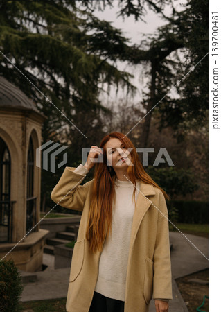 red-haired girl with long hair stands in park near building on a cloudy day red-haired girl with long hair stands in park near building on a cloudy day 139700481