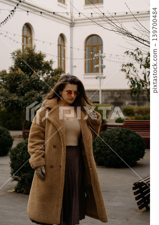 Woman walks in outdoor area wearing coat and sunglasses during daytime near a building Woman walks in outdoor area wearing coat and sunglasses during daytime near a building 139700484
