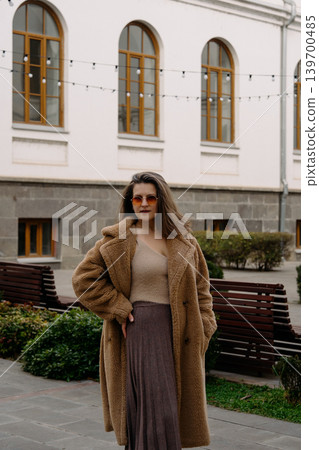 Woman standing in courtyard wearing coat and sunglasses during daytime in city Woman standing in courtyard wearing coat and sunglasses during daytime in city 139700485