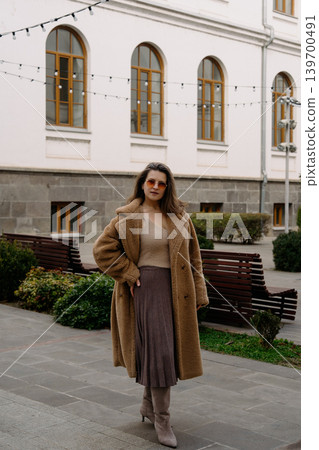 Woman stands in a coat and skirt on a city street near benches and buildings during the day Woman stands in a coat and skirt on a city street near benches and buildings during the day 139700491
