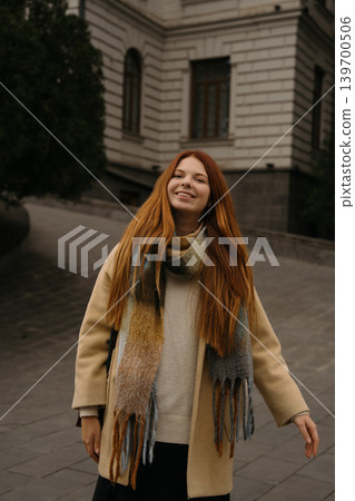 Smiling red-haired girl walks down a street in a city during daylight wearing a coat and scarf Smiling red-haired girl walks down a street in a city during daylight wearing a coat and scarf 139700506
