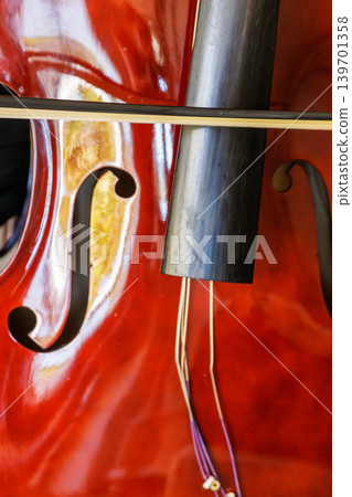 Close up of a glossy contrabass body with f hole, bow, tailpiece, and loose detached strings hanging over the varnished wood. Close up of a glossy contrabass body with f hole, bow, tailpiece, and loose detached strings hanging over the varnished wood. 139701358